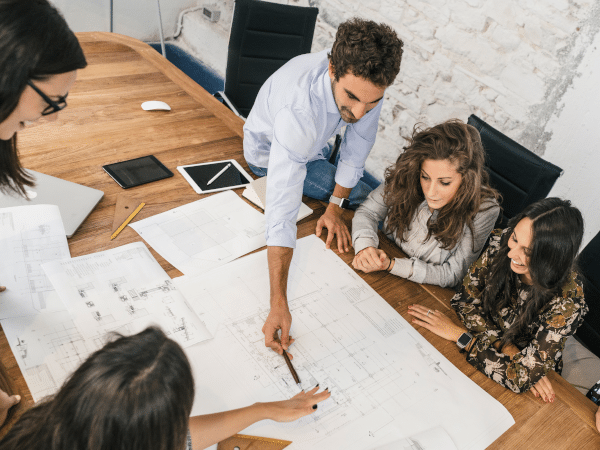 Equipe de jovens profissionais discutindo plantas e desenhos técnicos (impressão de projetos) numa mesa de reunião.